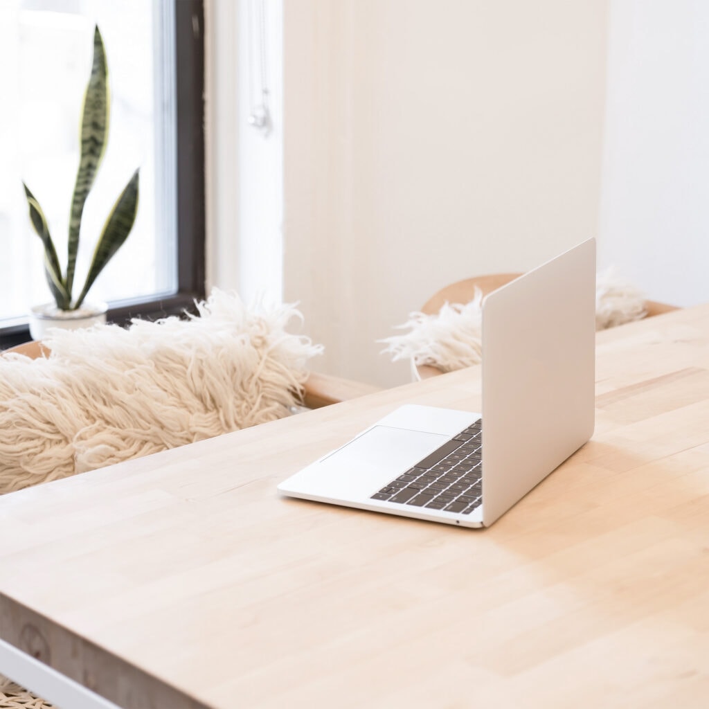 Rose gold laptop on a light wooden desk with a fluffy pillow on the chair. A workspace for a blogger