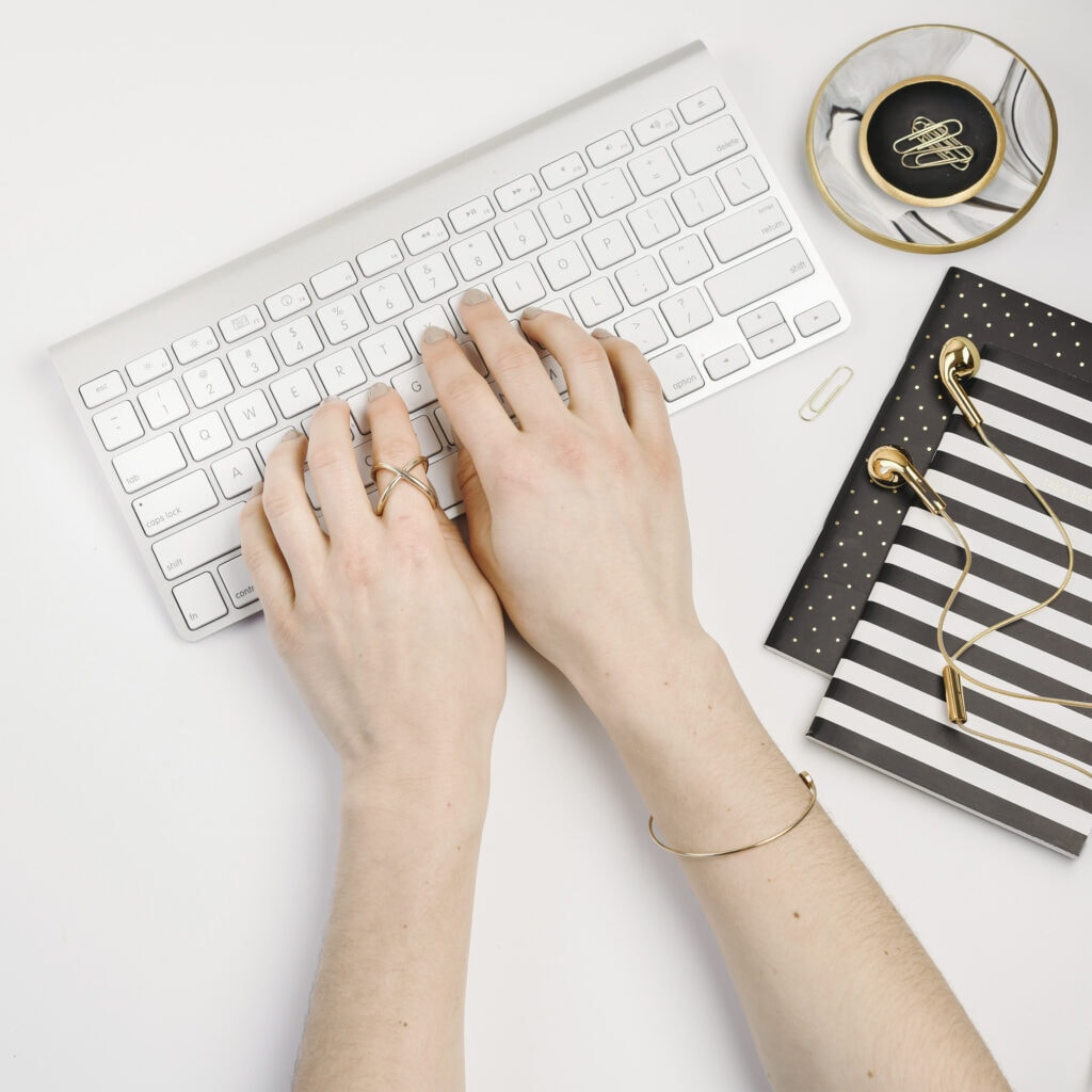 Woman's hands typing on an apple Bluetooth keyboard beside two notebooks. She is working on her SEO blog